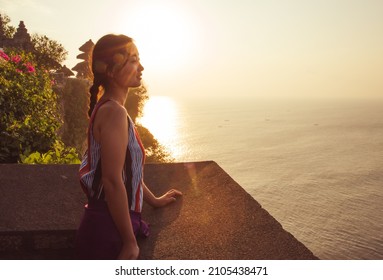Bali - Indonesia - 10.21.2015: Asian young female with braided dark hair watching the sea at sunset - golden hour at Uluwatu Temple - light beam