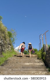 BALI, INDONESIA - Oct 6, 2016: Young Sporty Young Caucasian Couple With Hat And Surf Board Walking Up The Stairs. Blue Sky With Big Moon.