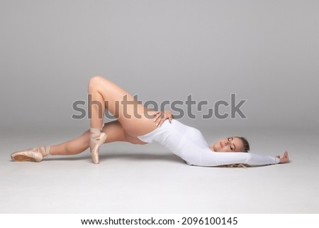 Ballerina, blonde with curly hair posing in a white leotard, on pointe shoes lying on the floor of the studio. Gray background. Flash.
