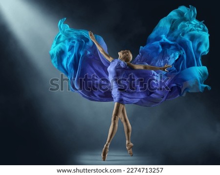 Ballerina in Purple Chiffon Dress on Stage Light Beam. Ballet Dancer in Silk Fantasy Blue Gown. Woman dancing with flying Cyan Fabric as Wings over Dark Background