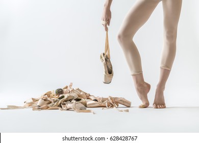 Ballerina stands on the toes and holds a pointe shoe on the ribbon on the light background in the studio. On the left side there is a pile of beige ballet shoes. Closeup. Horizontal.