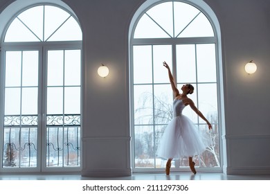 ballerina in white skirt and leotard on pointe shoes, against background of large windows with arches