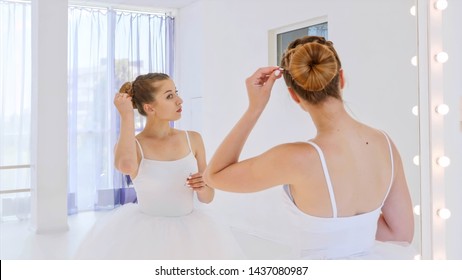 Ballerina in white tutu does her hair standing in front of the mirror in theatre dance class. She fixes the hair pulled into a bun with bobby pins. Portrait shot in mirror reflection.