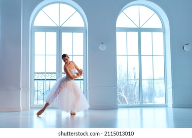 ballerina in white tutu and leotard on pointe shoes, against background of large windows with arches
