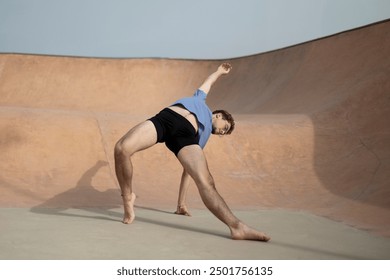 Ballet dancer in blue shirt and black shorts executes a dynamic side stretch in an empty skatepark, focusing on flexibility and grace on a sunny day. - Powered by Shutterstock