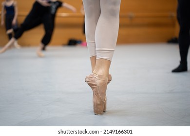 A Ballet Dancer Feet Doing Relevé. The Dancer Is Wearing Pointe Shoes And Cut Tights At The Level Of The Ankles. At The Background, Professional Dancers Training Hard.