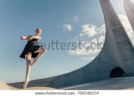 Ballet dancer practicing dance moves outdoors with blue sky and monument in the background. Female dancer balancing on one toe in pointe shoes on a rock.