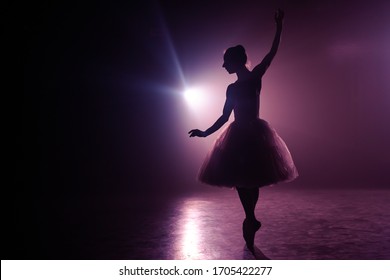 Ballet Dancer In Tutu Performing, Jumping On Stage. Ballerina Practices On Floor In Dark Studio With Smoke. Violet Light.