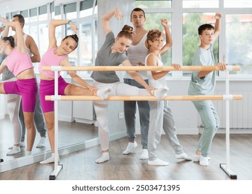 Ballet school students prepare for classes, near ballet bare stretch their leg muscles under middle-aged teacher guidance. Ballet class, studio, training of theatrical dance artists