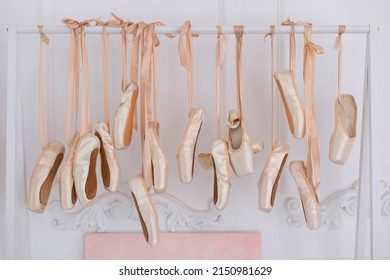 Ballet Shoes Hang On Bar In Room. Concept Of Dance, Ballet School, Ballerinas Clothes. Many Hanging Ballet Shoes On White Wall Background In Studio. New Pointe Shoes With Satin Ribbons Hanging On Ran