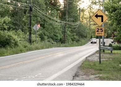 Baltimore County, MD US - May 20, 2021: Diamond Shaped Yellow Left Turn Arrow And 25 Mile Per Hour Speed Limit Signs Stacked Vertically On A Country Road  With A Car Approaching On The Other Side