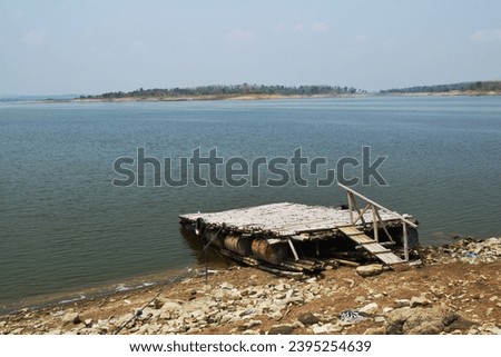 A bamboo boat dock is floating on the edge of the reservoir with ballast from barrels used against a background of shadows of the edges of the reservoir at a far.