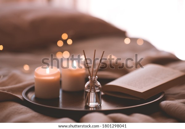 Bamboo sticks in bottle with\
scented candles and open book on wooden tray in bed closeup. Home\
aroma. 