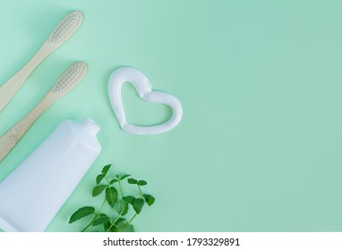 Bamboo Toothbrushes, White Tube Of Toothpaste, Mint Leaves On Light Green Background. Heart Shape Created From Tooth Paste. Top View, Copy Space