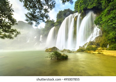 Ban Gioc Waterfall with lakeside tree foreground, smooth flow of water, creating in a waterfall down the white silk sheets high side mountains, sunny yellow iridescent color great lake