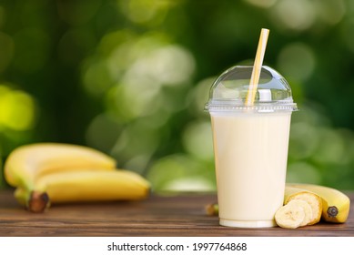 banana milkshake in disposable plastic glass with straw on wooden table outdoors