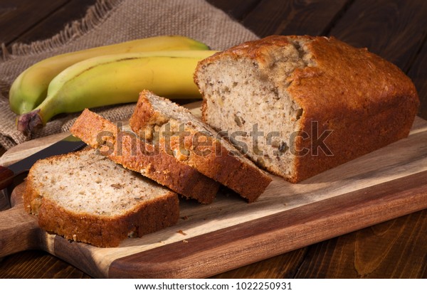 Banana nut sweet bread sliced on a wooden\
cutting board with bananas in\
background
