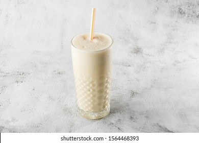 Banana Oats Smoothie Or Vanilla Milkshake In Glass On Bright Marble Background. Overhead View, Copy Space. Advertising For Milkshake Cafe Menu. Horizontal Photo.