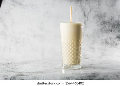 Banana Oats Smoothie Or Vanilla Milkshake In Glass On Bright Marble Background. Overhead View, Copy Space. Advertising For Milkshake Cafe Menu. Horizontal Photo.