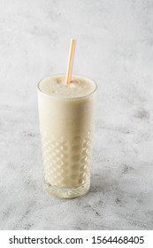 Banana Oats Smoothie Or Vanilla Milkshake In Glass On Bright Marble Background. Overhead View, Copy Space. Advertising For Milkshake Cafe Menu. Vertical Photo.