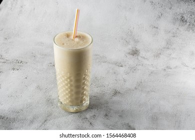 Banana Oats Smoothie Or Vanilla Milkshake In Glass On Bright Marble Background. Overhead View, Copy Space. Advertising For Milkshake Cafe Menu. Horizontal Photo.