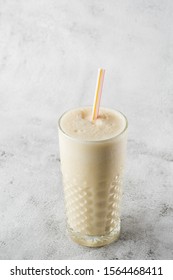 Banana Oats Smoothie Or Vanilla Milkshake In Glass On Bright Marble Background. Overhead View, Copy Space. Advertising For Milkshake Cafe Menu. Vertical Photo.