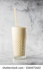 Banana Oats Smoothie Or Vanilla Milkshake In Glass On Bright Marble Background. Overhead View, Copy Space. Advertising For Milkshake Cafe Menu. Vertical Photo.