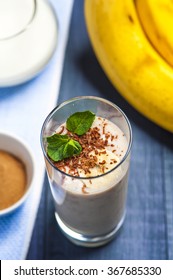 Banana Smoothie With Chocolate Flakes And Mint, Bananas, Cup Of Cinnamon And Yogurt On Blue Wooden Background. Conception Of Healthy Food. Focus On Mint And Banana Slice. Overhead View.