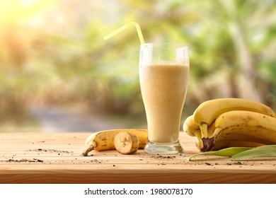 Banana smoothie in glass with straw on wooden table with fruit and plantation in the background. Front view.