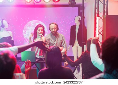 Band moving to rhythm while singing and mixing music on club stage illuminated with spotlights. Dj playing and singer with microphone dancing at discotheque in dark nightclub