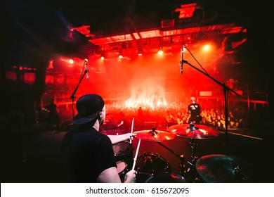 Band On Stage. Drummer Playing Drum Set At Concert On Stage. Music Show. Bright Scene Lighting In Club,drum Sticks In Hands.Fans Burn Red Flares At Rock Concert. View From The Stage To The Drummer. 