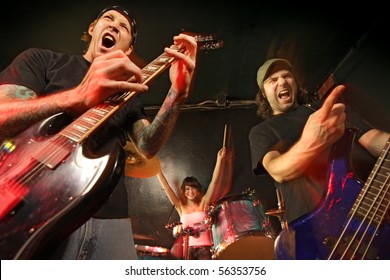 Band Playing On A Stage. Guitarist, Bassist And Female Drummer. Shot With Strobes And Slow Shutter Speed To Create Lighting Atmosphere And Blur Effects. Slight Motion Blur On Performers.