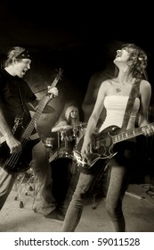 Band Playing On A Stage. Male Bassist With Female Guitarist And Drummer. Shot With Strobes And Slow Shutter Speed To Create Lighting Atmosphere And Blur Effects. Slight Motion Blur On Performers.