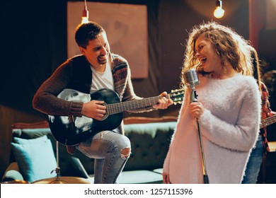 Band Practice For The Show. Woman With Curly Hair Holding Microphone And Singing While Man In Background Playing Acoustic Guitar. Home Studio Interior.