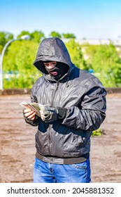 A Bandit, A Robber, A Thief, A Criminal, A Terrorist Stands With Surprised, Bulging Eyes In A Mask And Counts The Looted, Criminally Taken Money, Bills