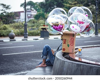 Bandung, Indonesia - May 4, 2021: A Man Selling Balloons Sitting With One Leg Up On The Side Of The Road While Resting