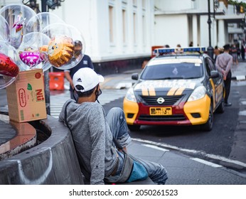 Bandung, Indonesia - May 4, 2021: A Man Selling Balloons Sitting With One Leg Up On The Side Of The Road In Front Of A Police Car While Resting