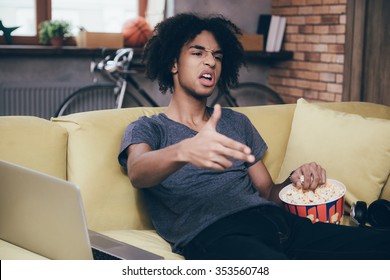 Bang! Young African Man Watching TV And Gesturingfinger Gun While Sitting With Bucket Of Popcorn On The Couch At Home