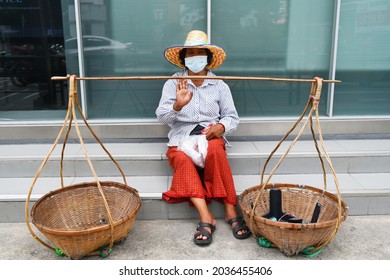 BANGKOK, THAILAND - AUGUST 2021: thai street seller with wooden baskets on pavement wait for customers. Asian market vendor on Sukhumvit road wearing farmer hat and face mask during COVID-19 lockdown.
