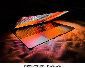 Bangkok, Thailand - May 3, 2021: Apple MacBook Retina Laptop Computer With Lid Half Open In Bed Sheets Lit With Warm Tone Vivid Desktop Screen Wallpaper In A Dark Room