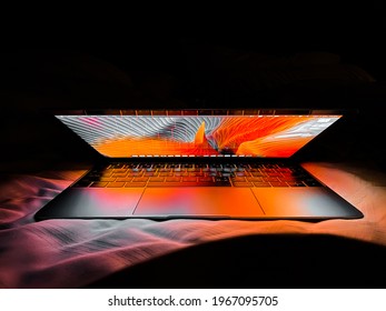 Bangkok, Thailand - May 3, 2021: Apple MacBook Retina Laptop Computer With Lid Half Open In Bed Sheets Lit With Warm Tone Vivid Desktop Screen Wallpaper In A Dark Room