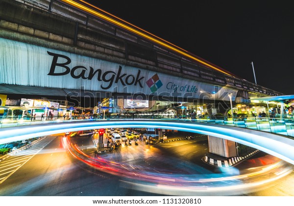 Bangkok, Thailand - Nov 25, 2017: Traffic light\
trail at road junction near MBK shopping center in Bangkok,\
Thailand. Tourism advertising logo on BTS sky train track,\
pedestrain walks on skywalk\
area