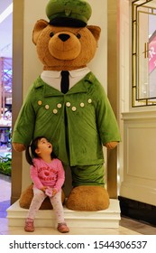 Bangkok / Thailand - October 12th, 2019: A Cute Thai Girl Sitting At The Foot Of A Big Harrods Teddy Bear With Green Jacket And Hat, Standing In Front Of The Store Inside Siam Paragon Shopping Center.