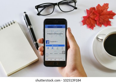 BANGKOK,THAILAND -January 14,2018: Hand holding Apple iPhone7 with Facebook application on the screen with Notebook, Coffee cup, Pen, Glasses and Maple leaf on White background