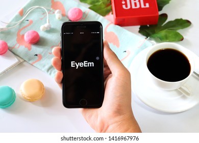 BANGKOK,THAILAND - June 2,2019:Hand Holding Apple IPhone  Opened EyeEm App On The Screen With Coffee Cup ,Loudspeaker ,Earphone ,Tropical Leave And Macaroon On White Background 