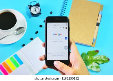 BANGKOK,THAILAND -September 23,2018: Hand holding Apple iPhone7 with Google application on the screen with Coffee cup, Notebook,clock, Tropical leaf , Color tag and Coffee beans on blue background