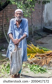 Bangladesh, Rangpur- September 07, 2021. An Old Famine-stricken Old Man Standing In Wearing Dirty Clothes With A Bag And A Stick In His Hand And Behind The Background Blur.