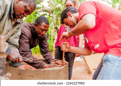 Bangoua, Cameroon - 08 august 2018: young european volunteer in african village building water well with spatula and concrete in collaboration with local old man