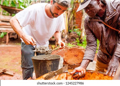 Bangoua, Cameroon - 08 August 2018: Young European Man In African Village Doing Manual Work Building Water Well With Concrete In Copperation With Local Old Man