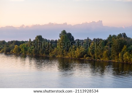 The bank of the Daugava River, overgrown with forest at sunset. Reflection of trees on the surface of the water. Beautiful big cloud of reconciliation. Natural landscape close up